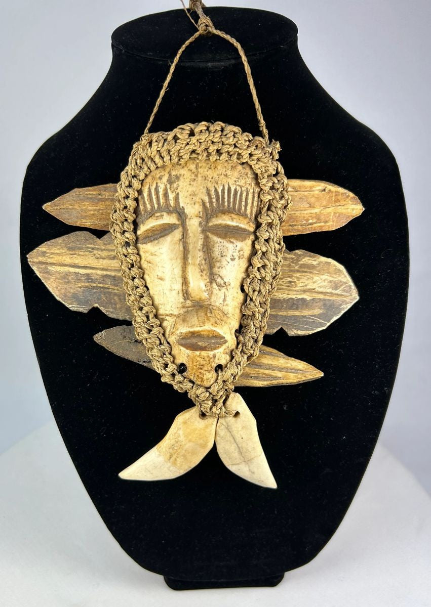 Mid-20th-century Lega tribal pendant mask from the Congo, carved bone face with braided fiber collar, wood backing, and hanging cord, displayed on a black stand against a white background.