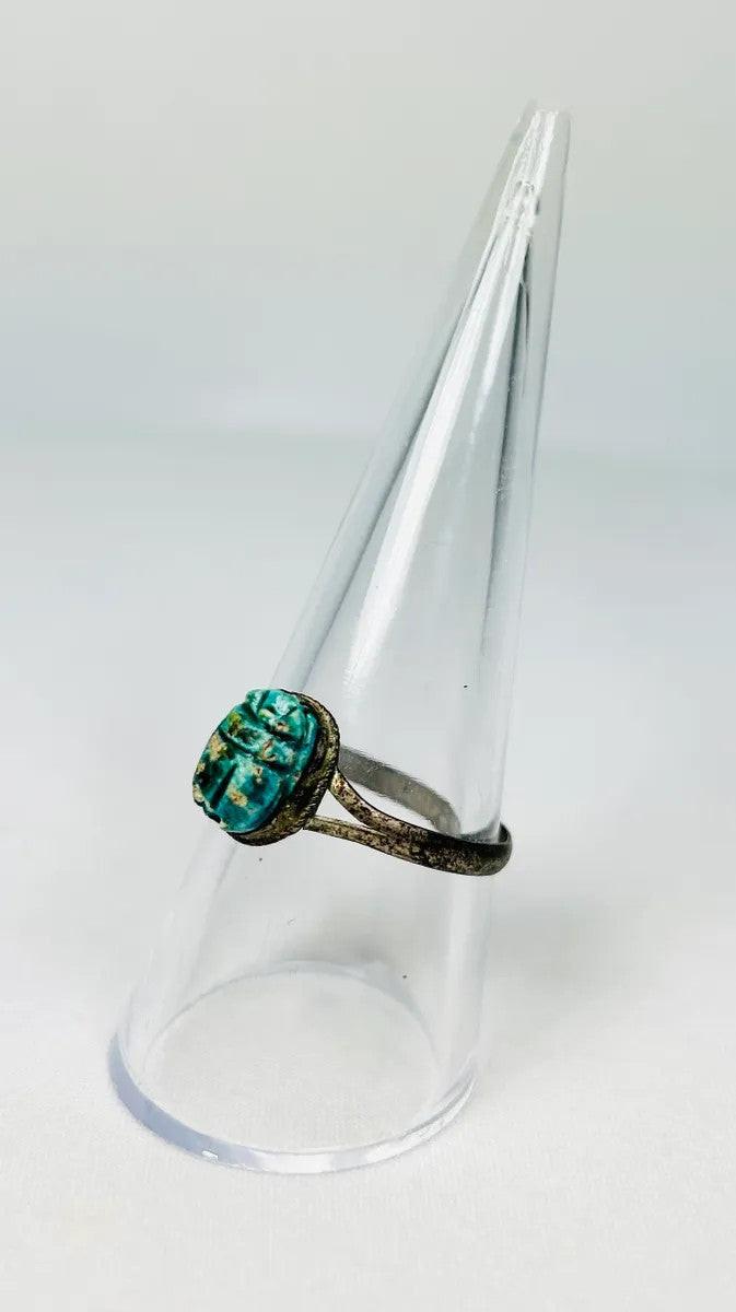 Ancient Egyptian scarab ring with turquoise-green faience scarab set in bronze band, displayed on a clear stand against a white background.