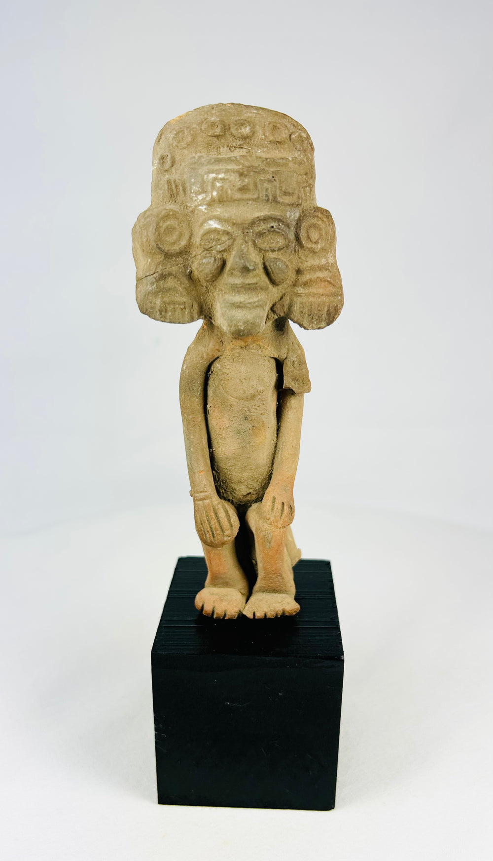 Mesoamerican Classic Period terracotta figure from Teotihuacan sphere with tall cylindrical headdress, incised roundels and step-fret band, large disk earspools, hands-on-knees posture, buff/orange clay with mineral accretions, mounted on modern black wood block, ca. 250–600 CE.