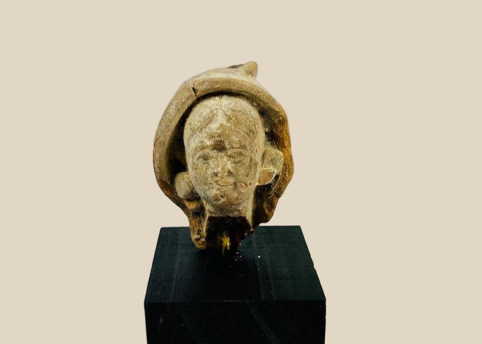 Terracotta head fragment from Classical Mediterranean, likely Greek or Roman, 1st Millennium BCE–CE. Features serene facial expression, partially preserved headdress, and natural weathered patina. Mounted on a custom stand, reflecting classical ideals of beauty, spirituality, and symbolic status in ancient art.