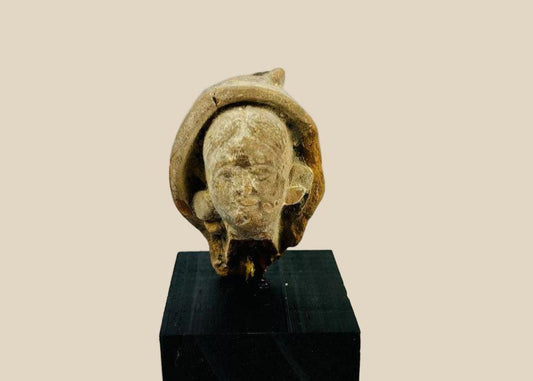 Terracotta head fragment from Classical Mediterranean, likely Greek or Roman, 1st Millennium BCE–CE. Features serene facial expression, partially preserved headdress, and natural weathered patina. Mounted on a custom stand, reflecting classical ideals of beauty, spirituality, and symbolic status in ancient art.