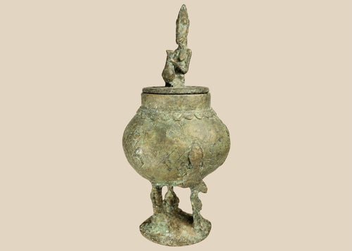 19th-century Dogon bronze container from Mali with bulbous body, engraved motifs, anthropomorphic legs, and a sculpted human figure on the lid, used in ritual and healing practices.