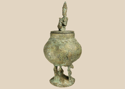 19th-century Dogon bronze container from Mali with bulbous body, engraved motifs, anthropomorphic legs, and a sculpted human figure on the lid, used in ritual and healing practices.