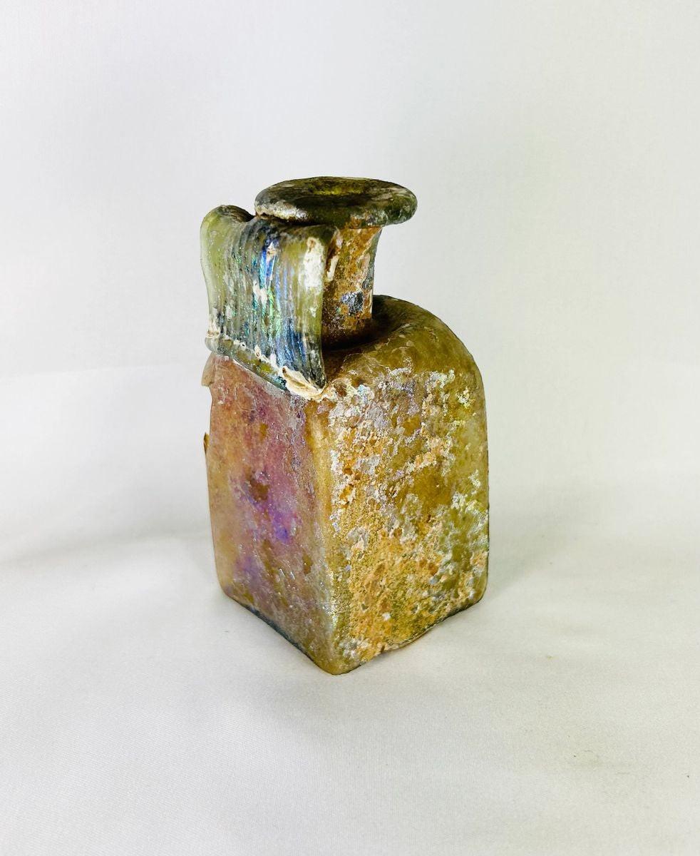 Early Imperial Roman mold-blown glass bottle from the 1st Century CE, likely Rhineland. Square translucent body with cylindrical neck, flaring folded rim, and wide ribbed “celery handle.” Base bears four-leaf maker’s mark. Developed iridescent patina reflects age and Roman glassmaking innovation, used for oils, perfumes, or wine.