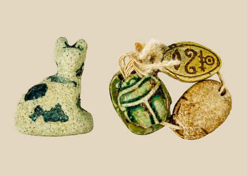 Ancient Egyptian faience cat amulet with traces of blue-green glaze, shown alongside three scarabs in varied materials, on a white background.