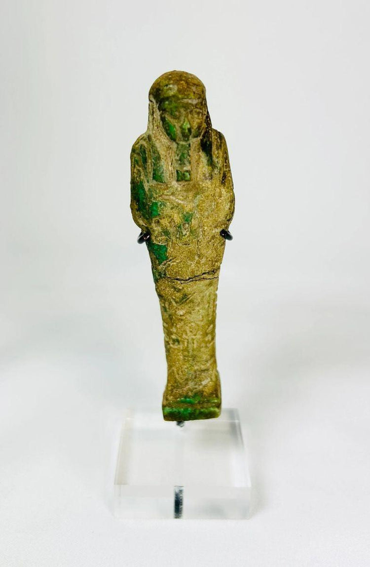 Ancient Egyptian green glazed faience Ushabti from the Late Period (664–332 BCE). Mummiform figure with tripartite wig, striated beard, and arms crossed holding hoe and pick. Hieroglyphic inscriptions adorn the body, symbolizing service in the afterlife. Reflects Egyptian funerary practices, craftsmanship, and spiritual beliefs.
