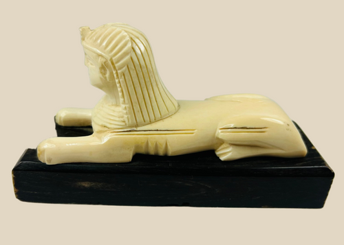 Early 20th century Egyptian revival Sphinx sculpture, reclining lion body with nemes headdress, hand-carved natural material mounted on black wooden base, decorative Egyptomania piece