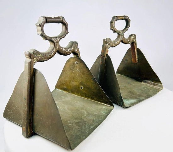 Antique pair of heavy brass equestrian stirrups with triangular footrests and intricately cast serpent motifs forming the handles, exhibiting detailed craftsmanship, natural patina, and symbolic protective design elements often associated with strength and guardianship in traditional horsemanship.