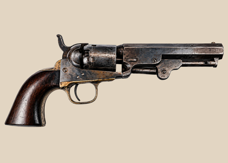 1859 Colt Model 1849 Pocket Revolver with aged steel, a brass trigger guard, and a walnut grip against a beige background.