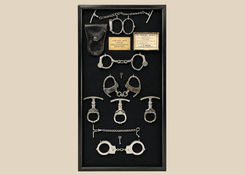 Framed shadow box display featuring a chronological collection of American and European handcuffs, leg irons, mechanical nippers, chain restraints, keys, and original leather pouches, mounted on black fabric with synthetic plaque, showcasing restraint devices from circa 1880s to 1950s.