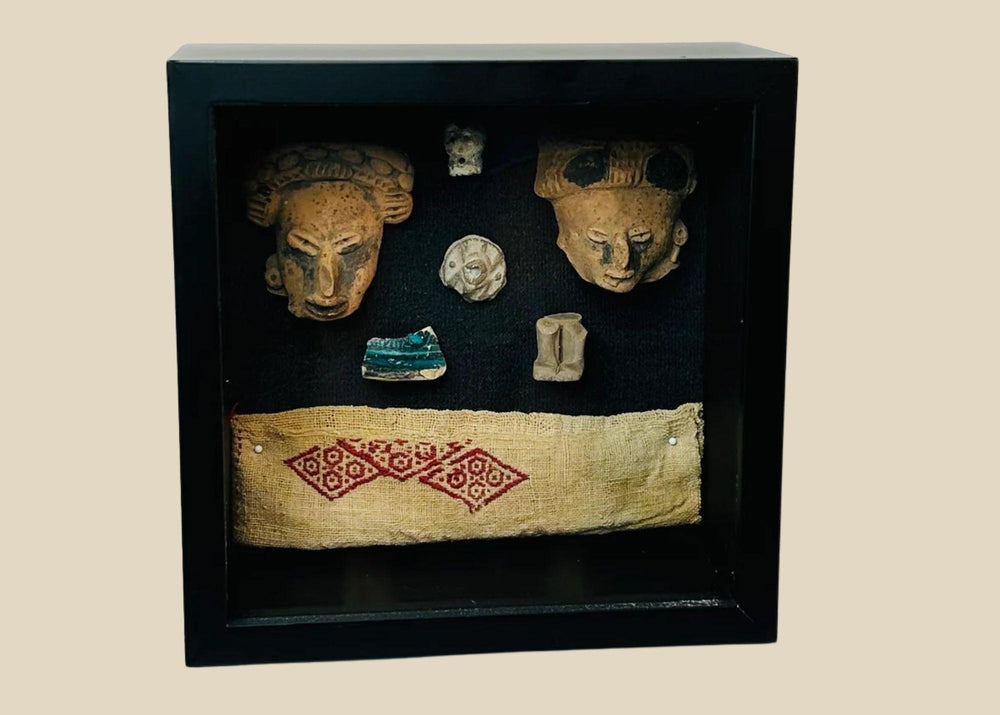 Framed display of Pre-Columbian artifacts including terracotta mask fragments, small figurines, carved ornaments, and a woven textile with red geometric pattern, set in a black shadowbox on a white background.