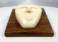 Abstract tribal-inspired bone face carving with closed eyes and simplified features, presented on a polished hardwood plaque.
