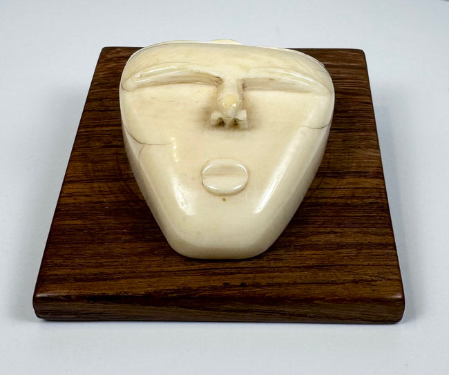 Vintage bone relief carving of a human spirit face with closed eyes and triangular form, mounted on a polished wooden plaque.