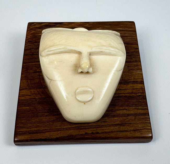 Mid-20th century bone carving depicting a stylized spirit face with an elongated nose, rounded lips, and smooth ivory-toned surface displayed on a wooden plaque.