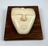 Mid-20th century bone carving depicting a stylized spirit face with an elongated nose, rounded lips, and smooth ivory-toned surface displayed on a wooden plaque.