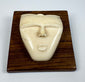 Mid-20th century bone carving depicting a stylized spirit face with an elongated nose, rounded lips, and smooth ivory-toned surface displayed on a wooden plaque.