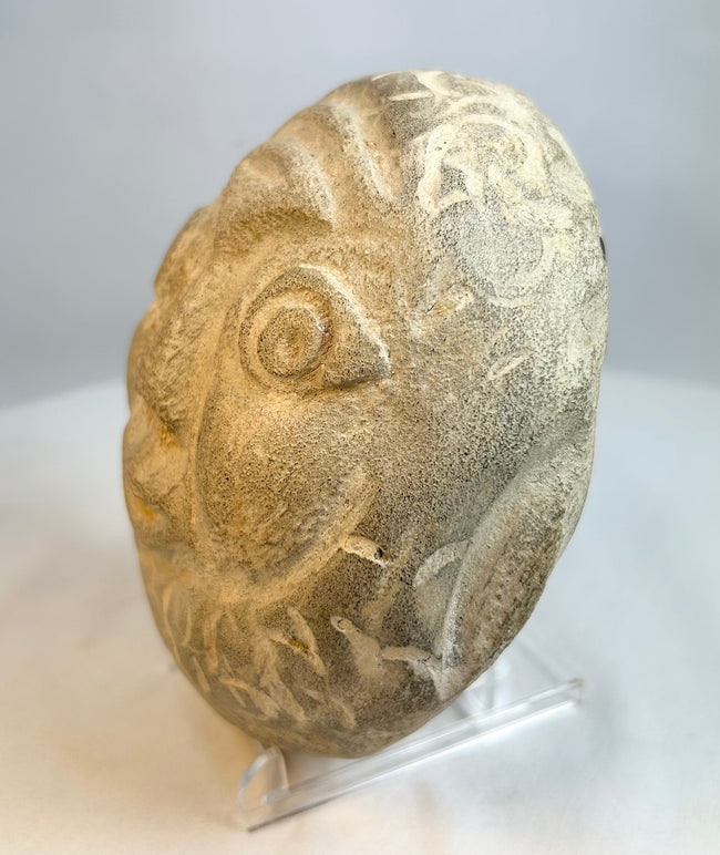 Rounded stone face sculpture displayed on clear stand with natural patina