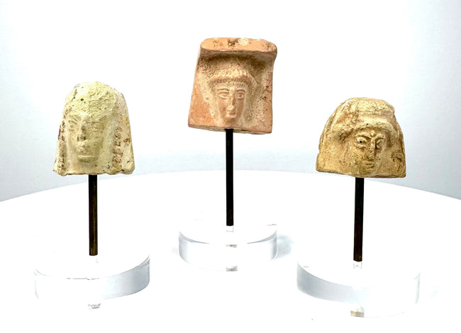 Collection of three ancient Greek female terracotta head fragments with stylized facial features, contrasting earthenware surfaces, and acrylic display stands.
