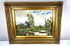 Early 20th-century landscape oil painting with a reddish-brown clothed figure in a wooden rowing boat surrounded by trees and grassy riverbanks.