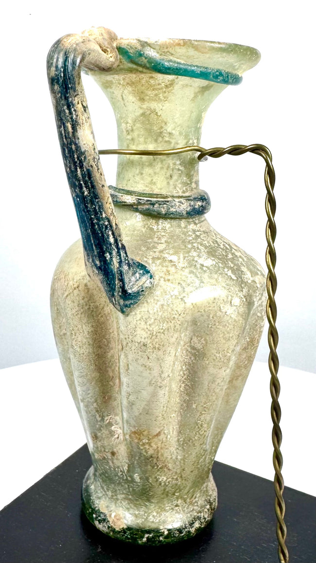 Ancient Roman glass pouring vessel with a gracefully curved handle, applied glass collar, and richly textured archaeological surface deposits.