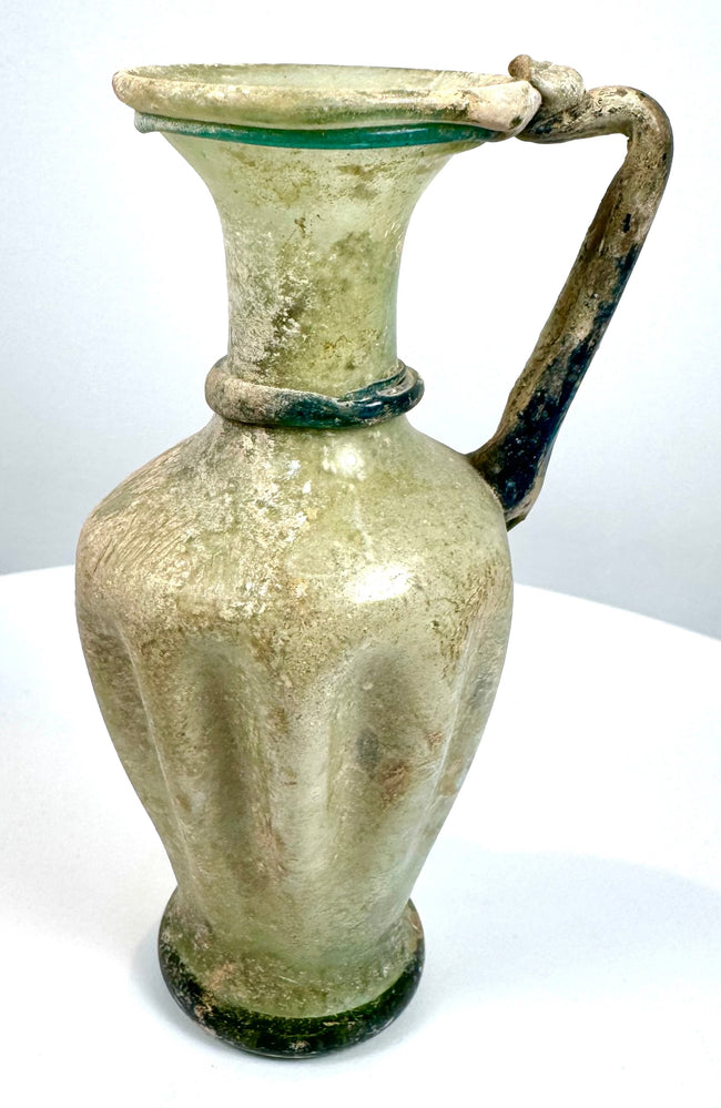 Ancient Eastern Mediterranean glass jug displaying textured vertical ribs, blue-green decorative glass threads, and natural surface deterioration.