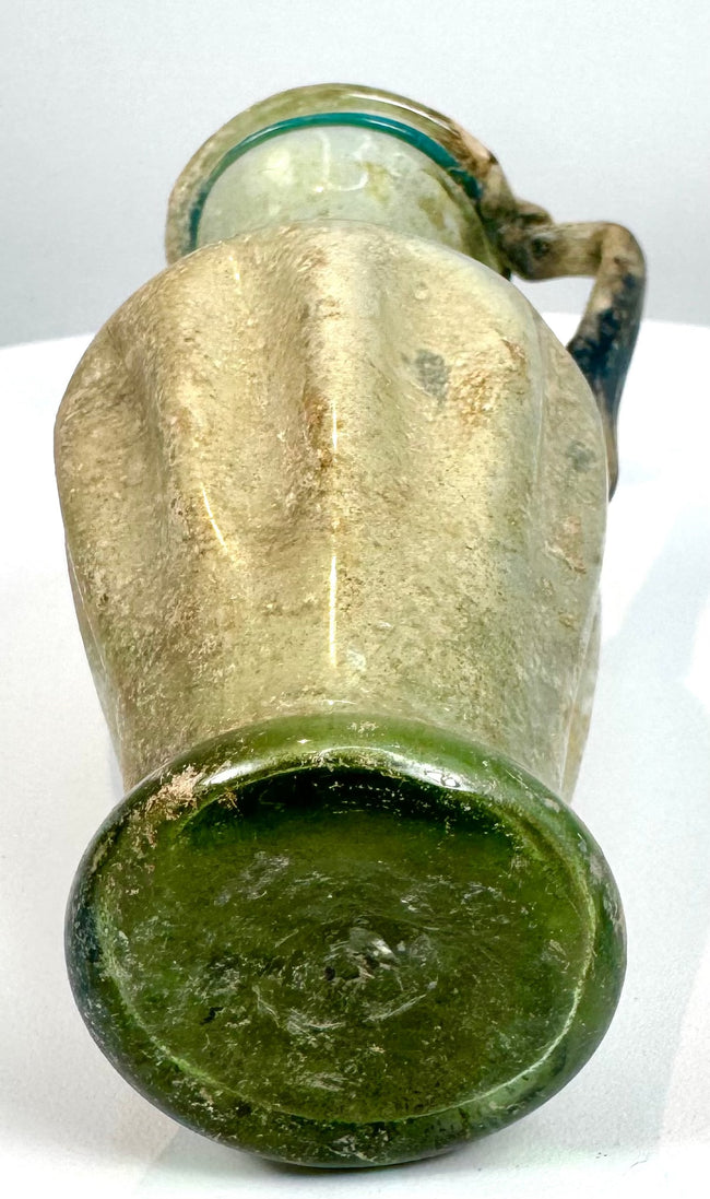 Ancient Roman–Byzantine glass jug, circa 4th–5th century CE, featuring pale green glass, extensive archaeological patination, and historical 1969 provenance.