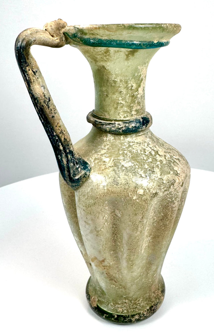 Ancient Roman ribbed glass jug exhibiting traditional pulled glass handle construction and applied decorative elements from the 4th–5th century CE.