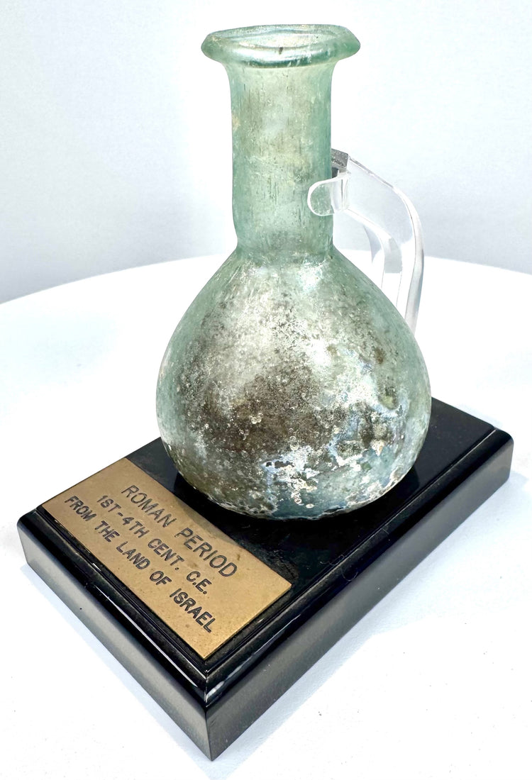 Ancient Roman glass perfume vessel mounted on a black display base with a historical identification plaque attributing it to the Roman Levant.