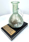 Ancient Roman glass perfume vessel mounted on a black display base with a historical identification plaque attributing it to the Roman Levant.