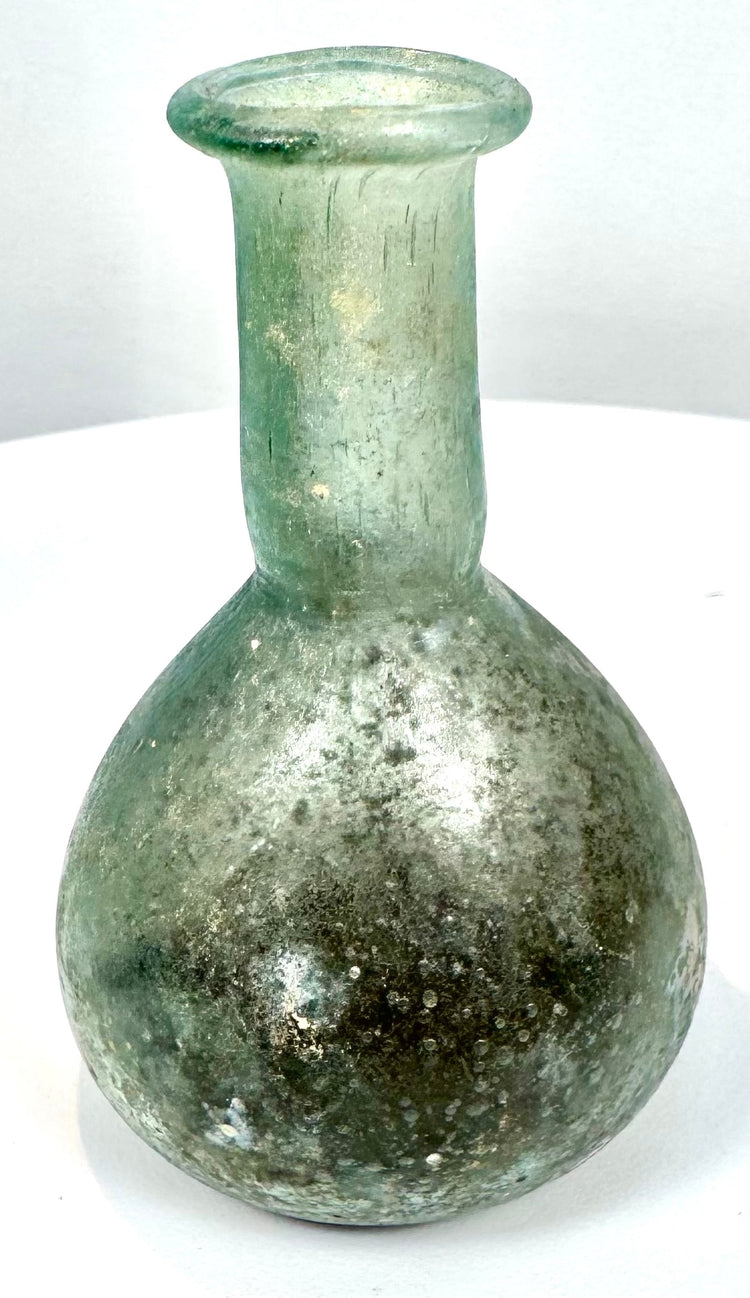 Ancient Roman pale green glass perfume bottle with mottled mineral deposits, localized iridescence, and a modern museum-style presentation base.