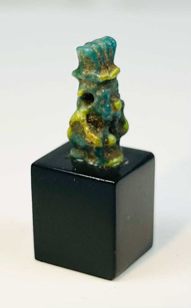 Ancient Egyptian Bes amulet in blue-green faience mounted on display stand.