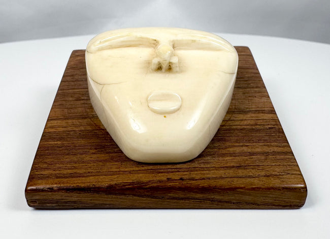 Abstract tribal-inspired bone face carving with closed eyes and simplified features, presented on a polished hardwood plaque.