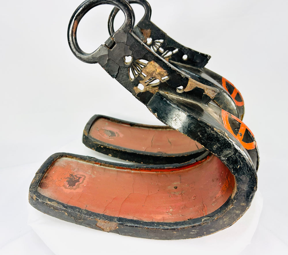 Pair of antique Japanese lacquered samurai stirrups with ornate cutout designs, red lacquer interiors, and cross-in-circle mon crests, Edo–Meiji period craftsmanship displayed on a white background.