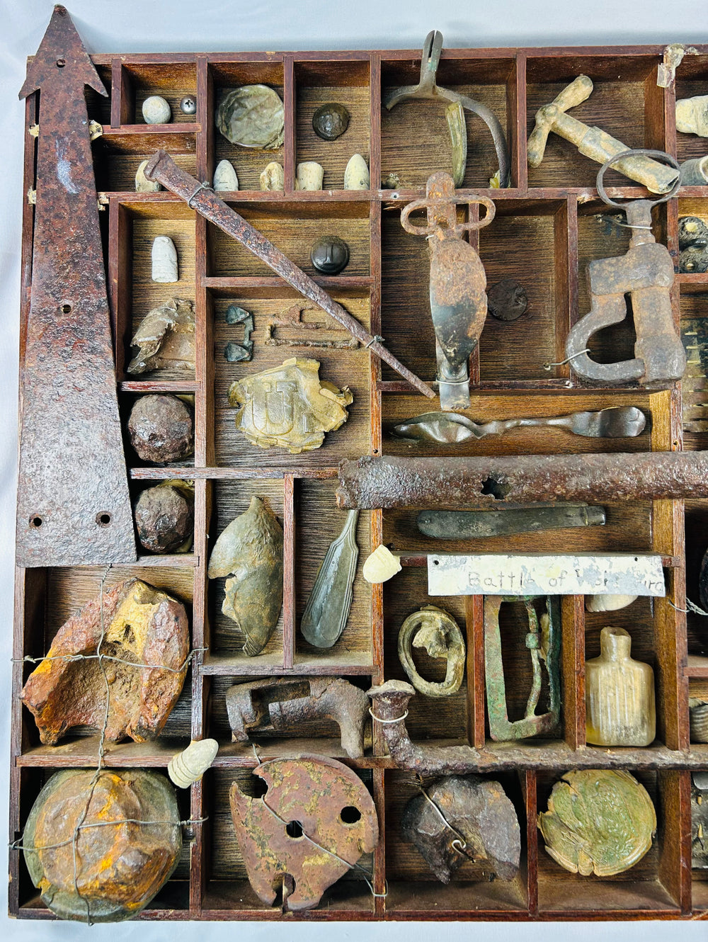 Excavated Civil War artifacts organized in a wooden display tray, featuring Minie balls, buckles, canteen spout, and iron camp tools.