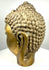 Antique-style cast-metal Buddha head featuring warm brass-like highlights, darker oxidation, and textured patination within the recessed sculptural details.