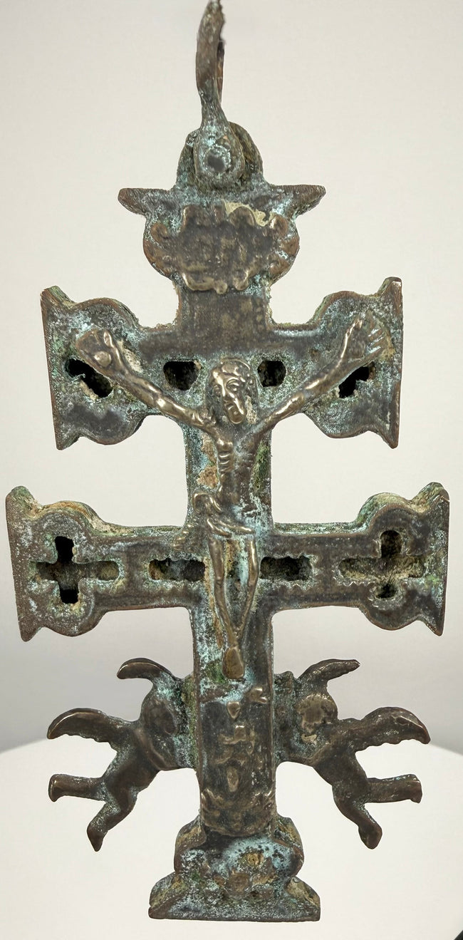 Spanish double-barred religious cross featuring sculpted religious figures, pierced openings, and a weathered surface with deep brown and green tones.