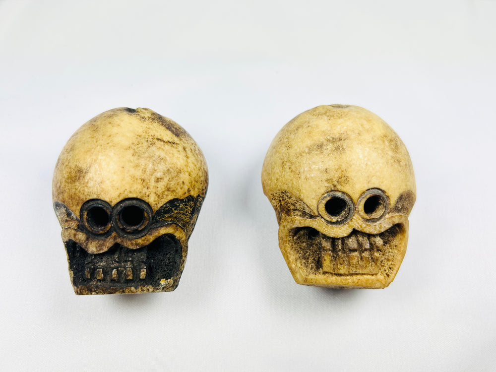 Front view of two Tibetan Citipati ritual beads carved from human femur bone, showing skull faces and suspension holes on a white background.