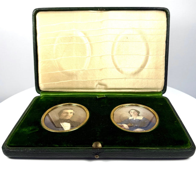 Front view of Victorian portrait miniatures depicting a gentleman in a dark formal coat and a woman in a blue gown, mounted in deep green velvet.