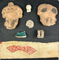 Pre-Columbian shadowbox with terracotta fragments, figurines, and red patterned textile on white background