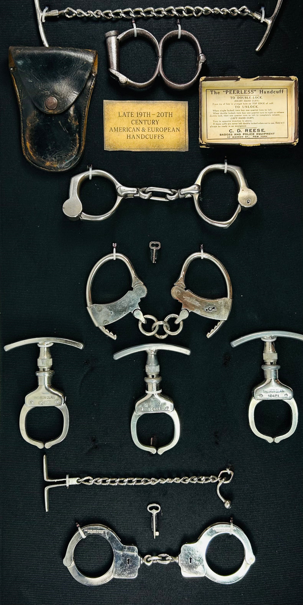 Framed collection of antique handcuffs, leg irons, and restraint devices from the 19th–20th century displayed on black fabric with original keys and plaques.