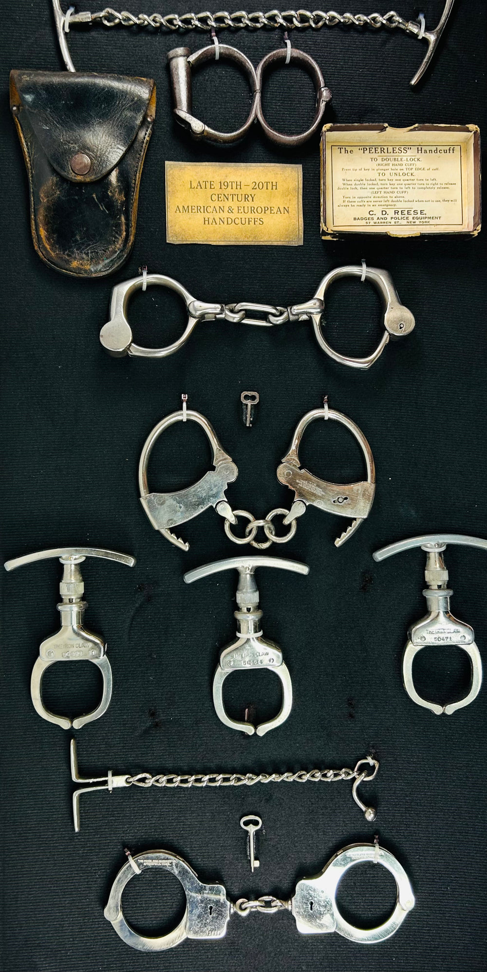 Antique handcuffs, keys, and leather pouch from a framed 19th–20th century restraint collection displayed on black fabric background.
