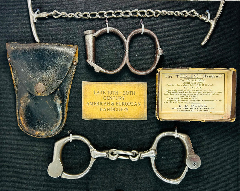 Close-up of late 19th–20th century American and European handcuffs with original leather pouch, identification plaques, and Peerless Handcuff label displayed on dark fabric.