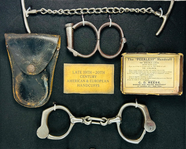 Close-up of late 19th–20th century American and European handcuffs with original leather pouch, identification plaques, and Peerless Handcuff label displayed on dark fabric.