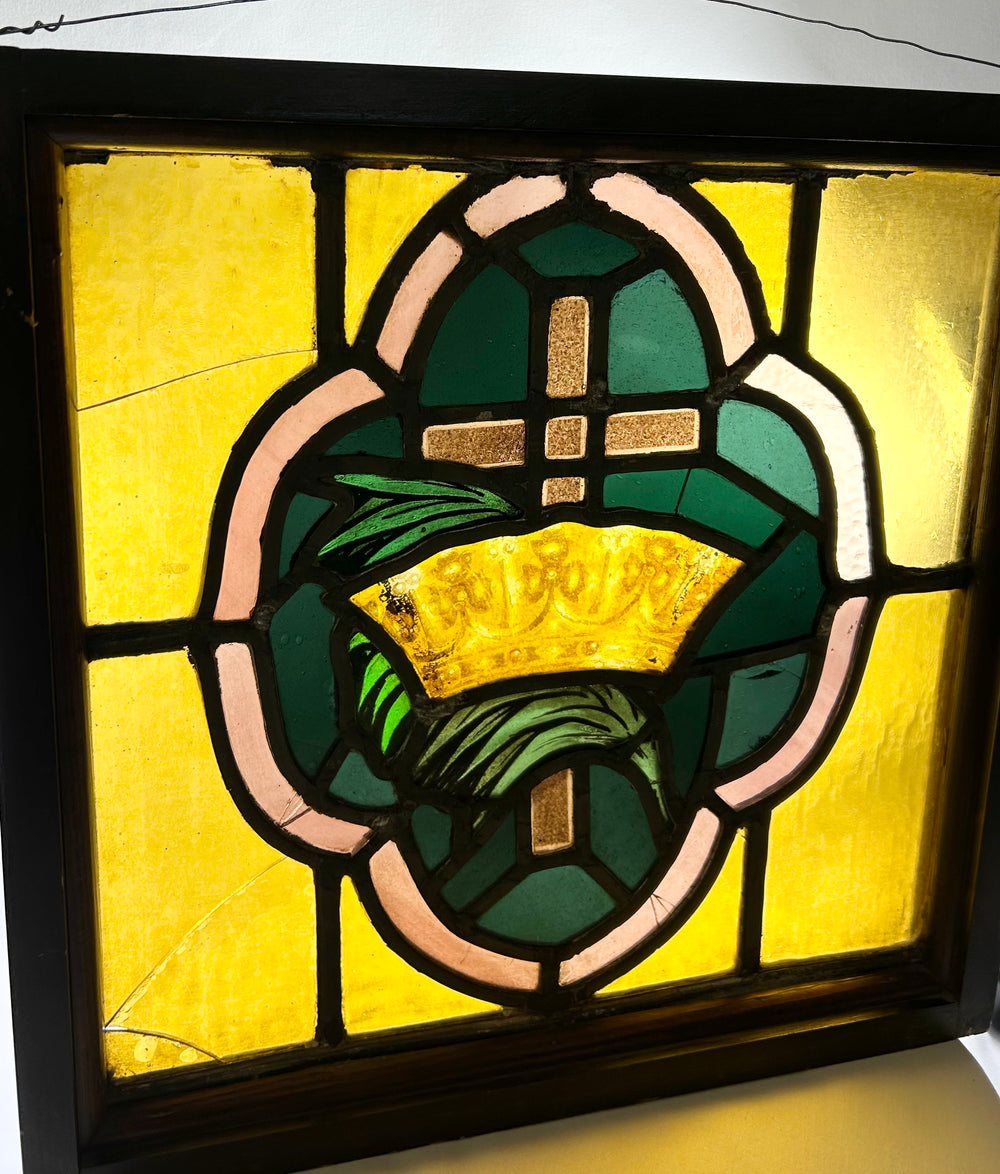 Wood-framed stained glass panel from early 20th century, featuring central cross and crown motif in amber, green, and pink, 16 inches square.
