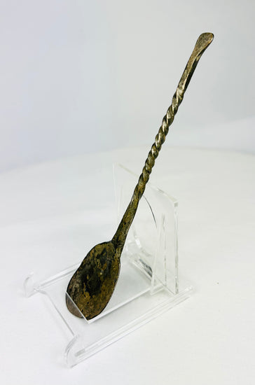 Low left angle of Roman or Byzantine bronze spoon emphasizing bowl curvature, handle twist, and aged bronze sheen.