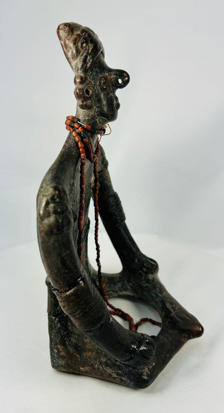 Profile view of ancient Mesoamerican shamanic figure with red beads and headdress, seated posture.