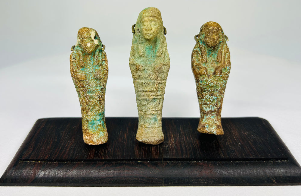 Front view of three ancient Egyptian faience ushabti figures on a dark wooden display stand with mineral-rich turquoise patina.