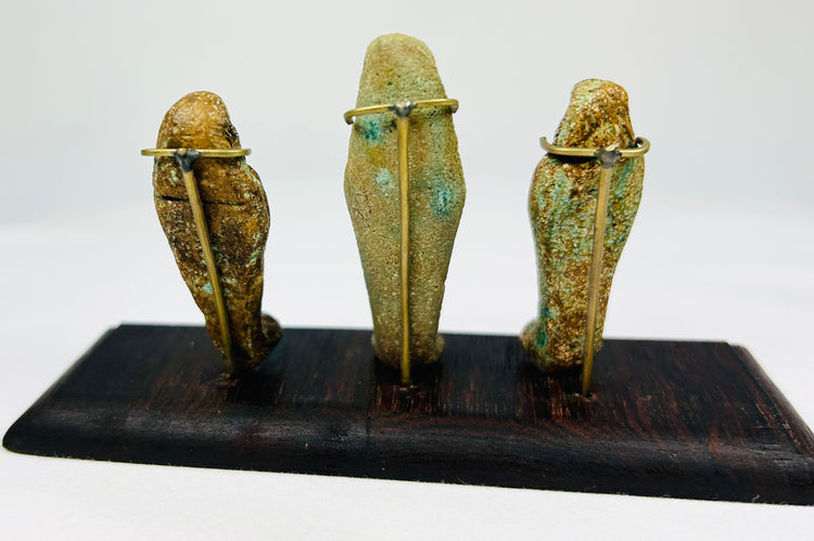 Rear view of three ancient Egyptian ushabti figures with burial patina and mounting pins displayed on a dark wood base.