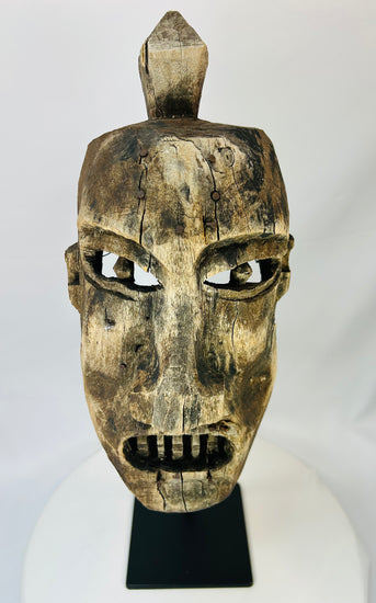 Front-facing image of a hand-carved tribal wooden mask mounted on a black stand.