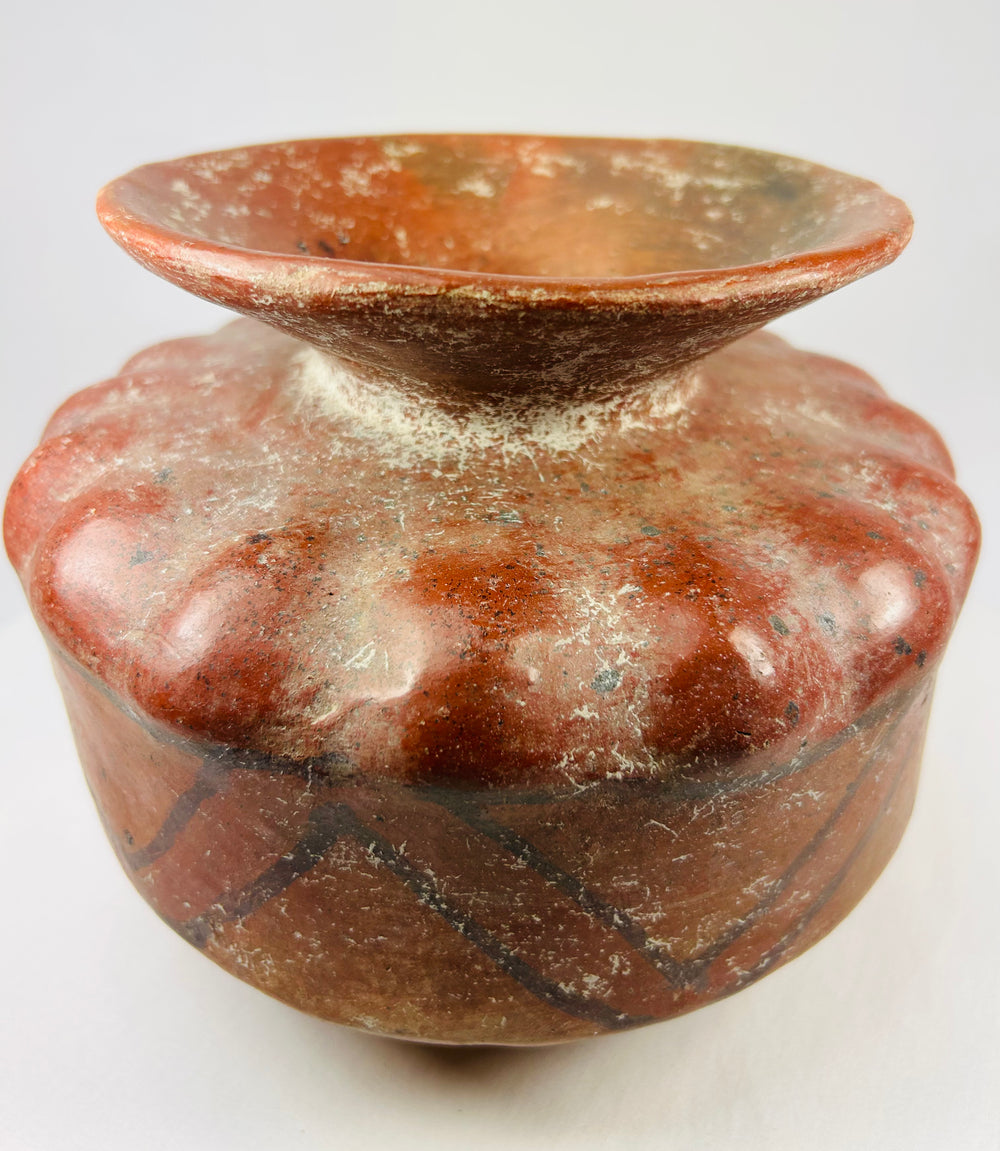 Pre-Columbian globular ceramic vessel with scalloped body and red-orange geometric painted designs on a white background.